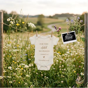 Puede incluir: Un body de bebé color crema con el texto "FOR THIS CHILD WE PRAYED" y la fecha "Noviembre 2026" cuelga de un tendedero con una imagen de ultrasonido. El fondo es un campo de flores silvestres.