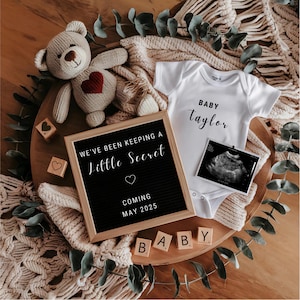 May include: A black letter board with the text "We've been keeping a little secret coming May 2025" and a white heart. A white onesie with the text "Baby Taylor" and a black and white ultrasound image. A knitted teddy bear with a red heart on its chest. Wooden blocks spelling "BABY".
