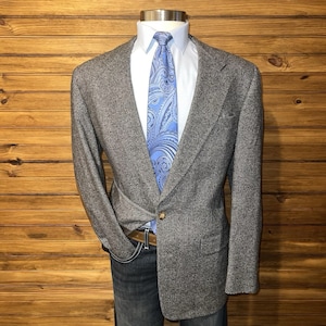 Può includere: Un blazer grigio in tweed con cravatta paisley azzurra e camicia bianca. Il blazer ha una chiusura a un bottone e un revers a lancia. L'outfit è abbinato a jeans blu e cintura marrone.