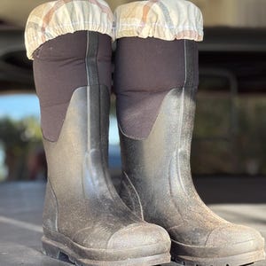 Puede incluir: Un par de botas de goma negras con cubiertas de tela de estampado marrón y blanco en la parte superior. Las botas están sucias y tienen una banda de rodadura gruesa.