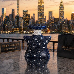 Pu&ograve; includere: Un flacone di profumo a forma di stella ricoperto di cristalli blu scuro e trasparenti, con un colletto argentato e un logo circolare con il testo Bond No. 9 New York. Il flacone &egrave; posizionato accanto a una borsa nera e un cartello Fifth Avenue New York, NY.