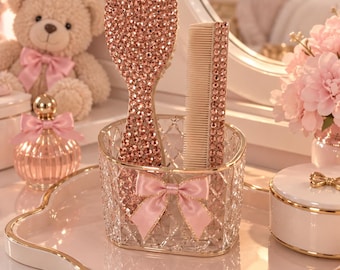 Può includere: Una spazzola per capelli e un pettine, entrambi ricoperti di strass color oro rosa, sono inseriti in un portaccessori in vetro trasparente con motivo a diamante. Il supporto è decorato sul davanti con un piccolo fiocco in raso rosa chiaro.