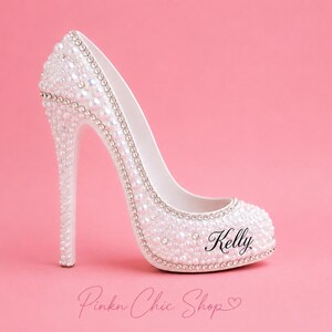 May include: A white high-heeled shoe figurine decorated with numerous small white pearls and clear rhinestones. The shoe features a thin stiletto heel and the name Kelly printed in black script on the side. The surface is covered in embellishments.