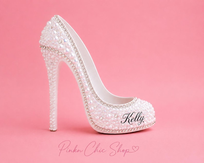 May include: A white high-heeled shoe figurine decorated with numerous small white pearls and clear rhinestones. The shoe features a thin stiletto heel and the name Kelly printed in black script on the side. The surface is covered in embellishments.