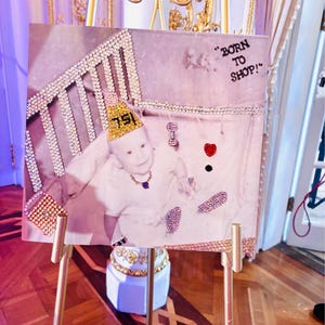 Pu&ograve; includere: Un cartello quadrato su un cavalletto dorato con la fotografia di un bambino che indossa un cappellino da festa giallo con il numero 75, una collana e un anello. L'immagine &egrave; decorata con strass che formano una culla, una borsa, un cuore e il testo BORN TO SHOP.