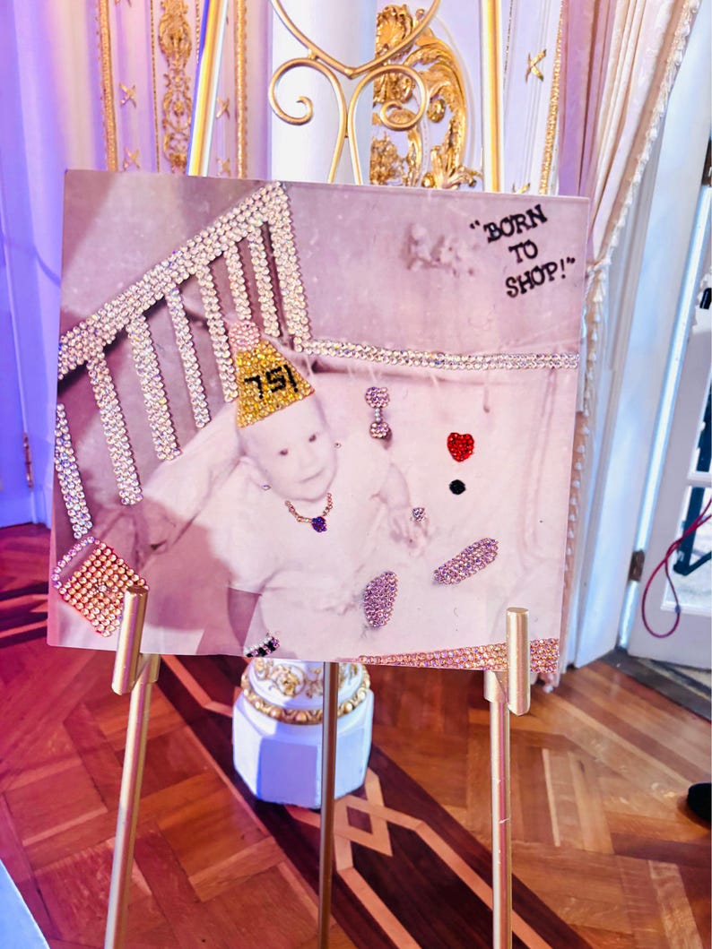 Pu&ograve; includere: Un cartello quadrato su un cavalletto dorato con la fotografia di un bambino che indossa un cappellino da festa giallo con il numero 75, una collana e un anello. L'immagine &egrave; decorata con strass che formano una culla, una borsa, un cuore e il testo BORN TO SHOP.