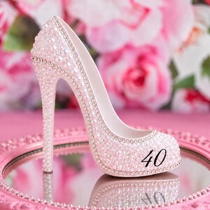 May include: A high-heeled shoe figurine covered in light pink pearls and small clear crystals. The side of the shoe features the black number 40. The shoe is positioned on a round, pink, ornate mirror tray with a beaded edge and a reflective surface.