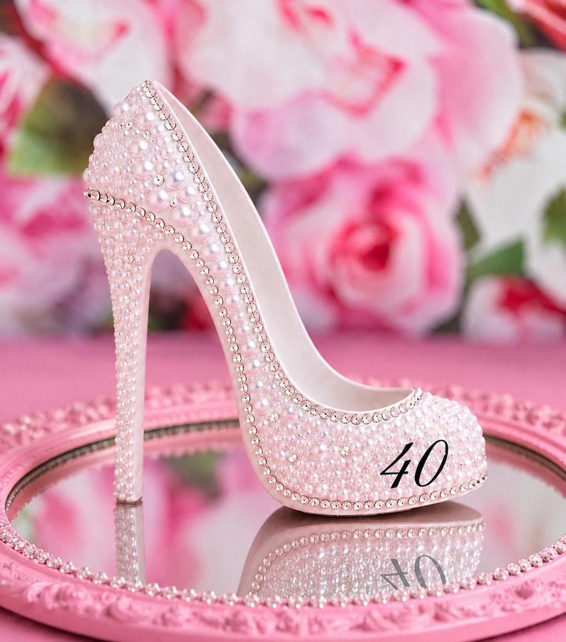 May include: A high-heeled shoe figurine covered in light pink pearls and small clear crystals. The side of the shoe features the black number 40. The shoe is positioned on a round, pink, ornate mirror tray with a beaded edge and a reflective surface.
