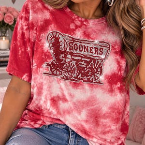 Pu&ograve; includere: Una t-shirt tie-dye rossa e bianca con una grafica centrale raffigurante un carro coperto e la scritta SOONERS, impreziosita da piccoli strass trasparenti. La maglia presenta un girocollo, maniche corte e un motivo marmorizzato su tutto il capo.