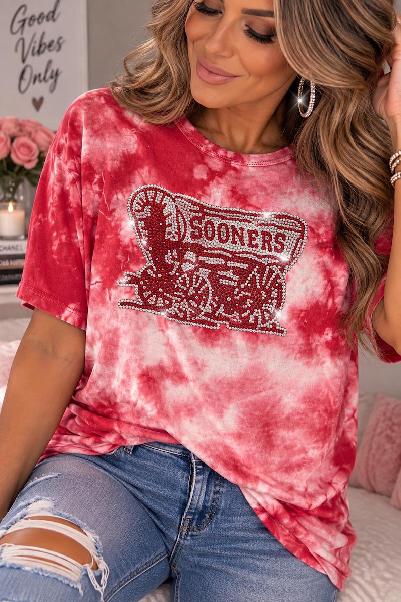 Pu&ograve; includere: Una t-shirt tie-dye rossa e bianca con una grafica centrale raffigurante un carro coperto e la scritta SOONERS, impreziosita da piccoli strass trasparenti. La maglia presenta un girocollo, maniche corte e un motivo marmorizzato su tutto il capo.