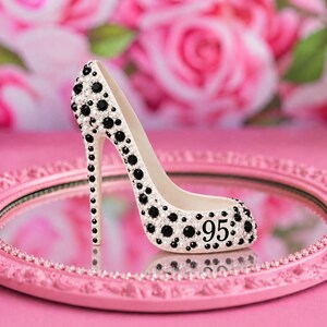 May include: A high-heeled shoe figurine decorated with white pearls and black rhinestones. The side of the shoe features the black printed number 95. The shoe is constructed with a pointed toe and a slender stiletto heel, covered in beads.