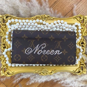 Pu&ograve; includere: Un portafoglio rettangolare marrone con l'iconico motivo monogramma marrone scuro e marrone chiaro. La parte anteriore del portafoglio &egrave; decorata con la parola "Queen" scritta con strass trasparenti e scintillanti in carattere corsivo.