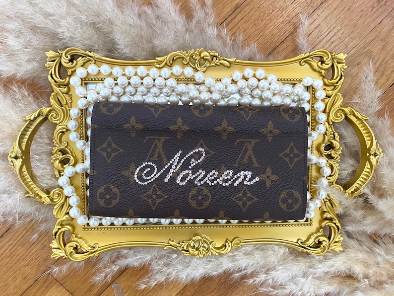 Pu&ograve; includere: Un portafoglio rettangolare marrone con l'iconico motivo monogramma marrone scuro e marrone chiaro. La parte anteriore del portafoglio &egrave; decorata con la parola "Queen" scritta con strass trasparenti e scintillanti in carattere corsivo.