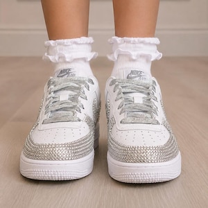 Pu&ograve; includere: Un paio di sneakers Nike Air bianche con decorazioni in strass argentati sulla punta e sui lati, abbinate a lacci glitterati argento e calzini bianchi con volant. Il logo Nike Air &egrave; visibile sulla linguetta di ogni scarpa.