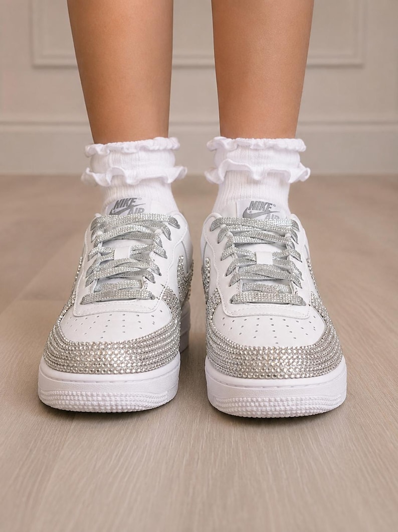 Pu&ograve; includere: Un paio di sneakers Nike Air bianche con decorazioni in strass argentati sulla punta e sui lati, abbinate a lacci glitterati argento e calzini bianchi con volant. Il logo Nike Air &egrave; visibile sulla linguetta di ogni scarpa.