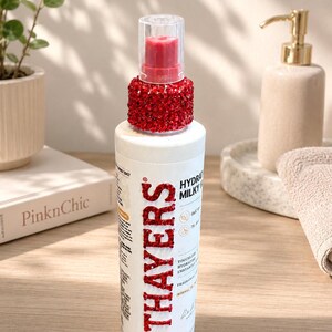 Pu&ograve; includere: Un flacone cilindrico bianco di tonico idratante Thayers con un tappo decorato con strass rossi e il nome del marchio Thayers scritto in strass rossi sulla parte anteriore. L'etichetta indica un volume di 130 ml.
