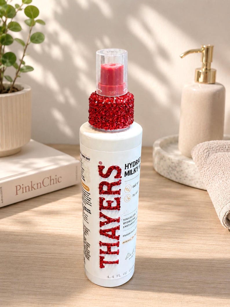 Pu&ograve; includere: Un flacone cilindrico bianco di tonico idratante Thayers con un tappo decorato con strass rossi e il nome del marchio Thayers scritto in strass rossi sulla parte anteriore. L'etichetta indica un volume di 130 ml.