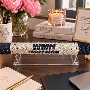 Pu&ograve; includere: Un bastone cilindrico poggiato su un supporto in acrilico trasparente, ricoperto di strass scintillanti trasparenti e blu scuro. Al centro presenta il testo WMN e LEGACY BATON in lettere blu scuro. Le estremit&agrave; sono rifinite con pietre blu scuro.