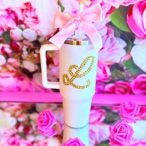 Pode incluir: Caneca branca com alça, adornada com uma letra "L" dourada com glitter. Um laço rosa está amarrado ao redor do canudo. O fundo apresenta um arranjo floral com rosas cor-de-rosa.