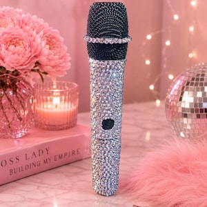 Pu&ograve; includere: Un microfono portatile con una testa a rete nera e un'impugnatura interamente ricoperta di strass trasparenti e riflettenti. Una fila di cristalli pi&ugrave; grandi circonda la base della testa del microfono e un tasto di accensione nero &egrave; visibile.