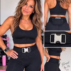 Pu&ograve; includere: Una cintura sportiva nera con una fibbia rettangolare tempestata di strass trasparenti. La cintura &egrave; indossata in vita sopra leggings neri e un reggiseno sportivo abbinato. &Egrave; visibile il testo Blinged by Me con un simbolo a cuore.