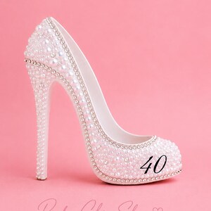 May include: A white high-heeled shoe decorated with numerous small white pearls and silver-toned rhinestones. The number 40 is printed in black on the side of the shoe. The text Pinkn Chic Shop with a heart symbol is visible at the bottom.