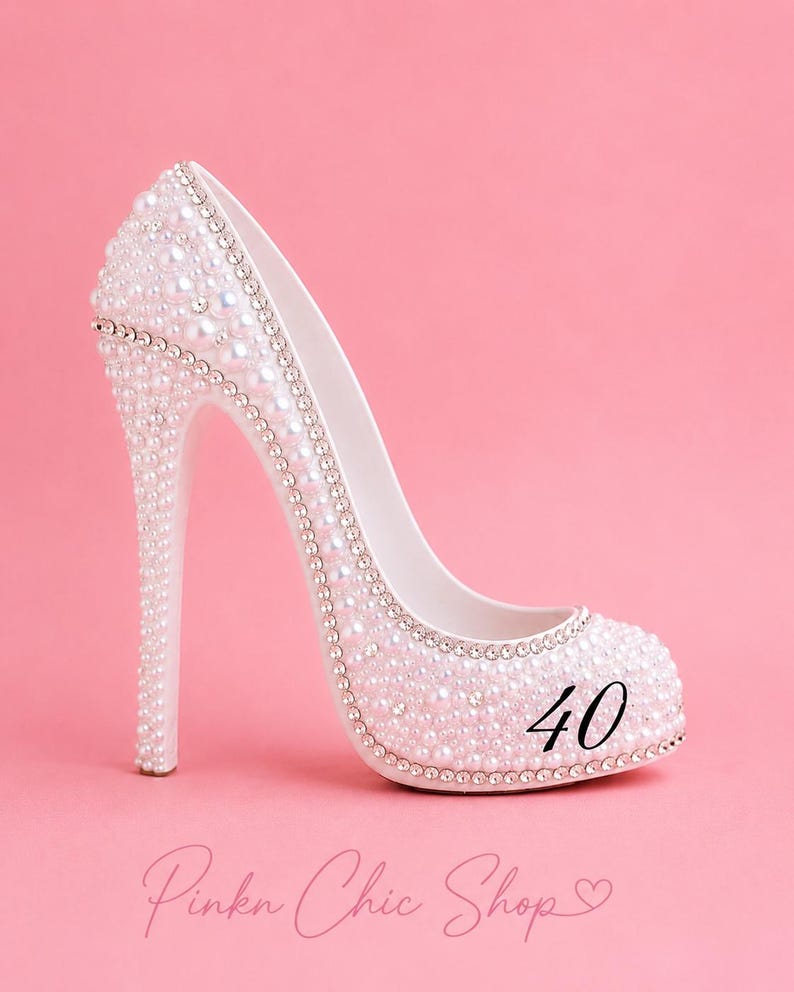 May include: A white high-heeled shoe decorated with numerous small white pearls and silver-toned rhinestones. The number 40 is printed in black on the side of the shoe. The text Pinkn Chic Shop with a heart symbol is visible at the bottom.