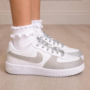 Pu&ograve; includere: Sneaker bianche con lacci glitterati argento e logo Nike swoosh laterale tempestato di strass argento. Anche la punta &egrave; ricoperta di strass argento. Abbinate a calzini bianchi alla caviglia con volant.