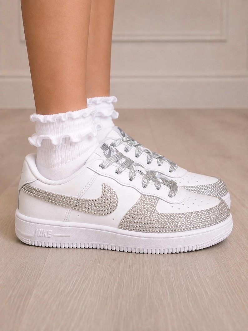 Pu&ograve; includere: Sneaker bianche con lacci glitterati argento e logo Nike swoosh laterale tempestato di strass argento. Anche la punta &egrave; ricoperta di strass argento. Abbinate a calzini bianchi alla caviglia con volant.
