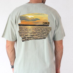 Puede incluir: Camiseta verde claro con un gráfico de puesta de sol sobre el agua en la espalda. El gráfico incluye un cielo amarillo y naranja, agua y el texto "7ONE8 OUTFITTERS". La camiseta tiene mangas cortas y cuello redondo.