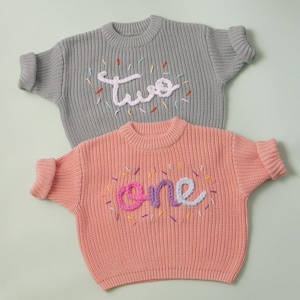 May include: Two knitted sweaters for babies, one grey with the word "two" embroidered in white yarn, and one pink with the word "one" embroidered in pink and purple yarn.