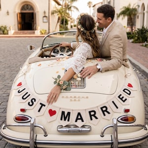 May include: A cream-coloured vintage Porsche 356 convertible decorated with a Just Married banner featuring red hearts and black lettering. The car is adorned with scattered flower petals, and the rear deck lid displays the Porsche 356 emblem.