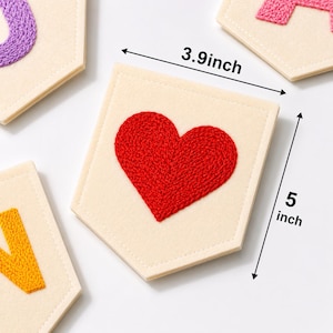 May include: A cream-coloured felt pennant featuring a red embroidered heart. The pennant is pentagonal in shape with stitched edges. Measurement arrows indicate a width of 3.9 inches and a height of 5 inches for the central decorative piece.
