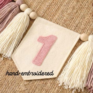 K&ouml;nnte beinhalten: Ein cremefarbenes Filzbanner mit einer handgestickten rosa Eins. Das Banner ist Teil einer Girlande mit cremefarbenen, rosa und malvenfarbenen Quasten, Holzperlen und dem Text "hand-embroidered".
