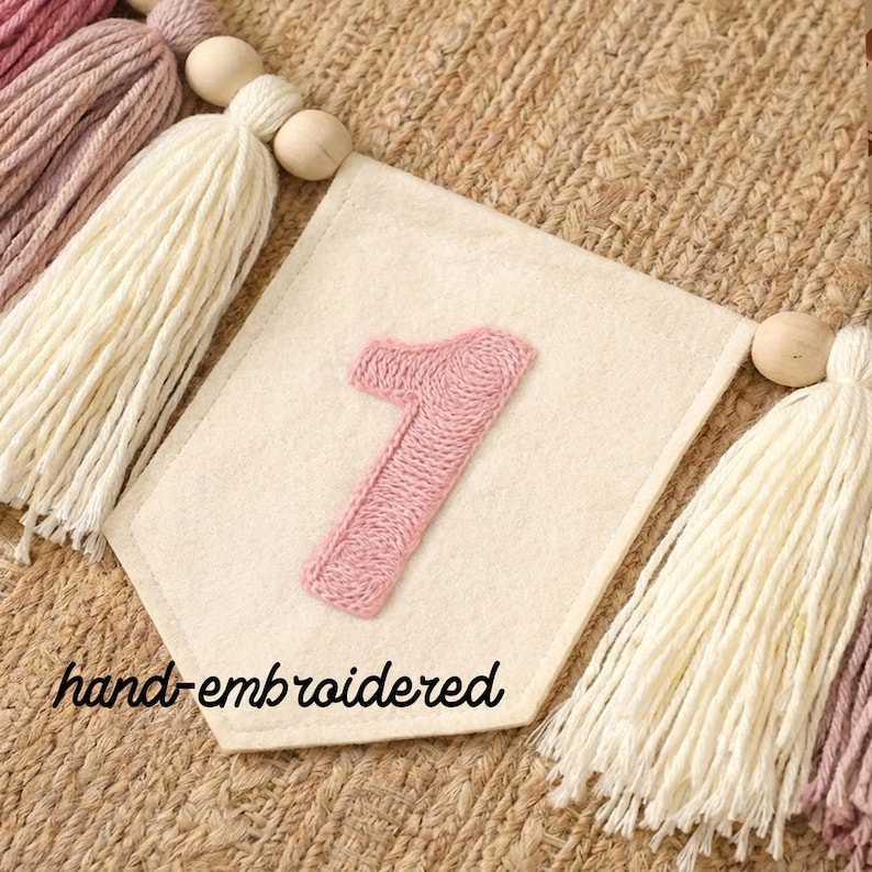 K&ouml;nnte beinhalten: Ein cremefarbenes Filzbanner mit einer handgestickten rosa Eins. Das Banner ist Teil einer Girlande mit cremefarbenen, rosa und malvenfarbenen Quasten, Holzperlen und dem Text "hand-embroidered".