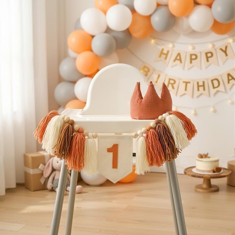 K&ouml;nnte beinhalten: Ein Hochstuhl, dekoriert f&uuml;r eine erste Geburtstagsfeier. Der Stuhl ist mit einer Quastengirlande, einem Banner mit der Zahl 1 und einer Krone geschm&uuml;ckt. Ballons in Orange, Wei&szlig; und Grau bilden den Hintergrund. Ein kleiner Kuchen steht auf einem Holzst&auml;nder.