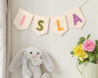 May include: A white fabric banner with the name "ISLA" spelled out in colorful felt letters. The letters are pink, green, orange, and purple. A gray stuffed bunny rabbit sits on a shelf below the banner.