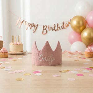 K&ouml;nnte beinhalten: Eine rosa Stoffkrone mit dem Namen "Emily" ist auf einem Holztisch platziert, umgeben von Konfetti. Ein kleiner Kuchen mit Kerzen, Cupcakes und Luftballons in Gold, Wei&szlig; und Rosa schaffen eine festliche Geburtstagsatmosph&auml;re.
