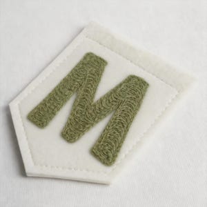 May include: A pentagonal patch featuring a white felt base with a stitched border and a large, textured letter M embroidered in a muted olive green thread. The design is centred on the white fabric, showcasing detailed needlework throughout.