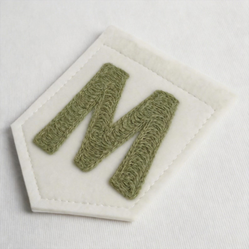 May include: A pentagonal patch featuring a white felt base with a stitched border and a large, textured letter M embroidered in a muted olive green thread. The design is centred on the white fabric, showcasing detailed needlework throughout.