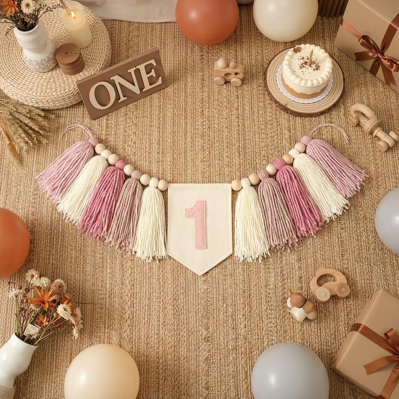 K&ouml;nnte beinhalten: Ein festliches Banner mit rosa, cremefarbenen und wei&szlig;en Quasten, Holzperlen und einem Wimpel mit der rosa Zahl "1". Das Banner ist Teil einer ersten Geburtstagsfeier mit Luftballons, Kuchen und Geschenken.
