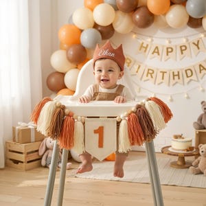 K&ouml;nnte beinhalten: Ein Baby mit einer Krone mit dem Namen "Ethan" sitzt in einem Hochstuhl, der mit einem "1"-Banner und einer Quasten-Girlande geschm&uuml;ckt ist. Im Hintergrund befinden sich Ballons in Braun-, Orange- und Cremet&ouml;nen sowie ein "Happy Birthday"-Banner.