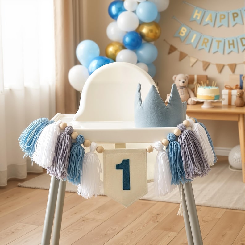 K&ouml;nnte beinhalten: Hochstuhl dekoriert f&uuml;r einen ersten Geburtstag. Er hat eine blaue Krone, ein Banner mit der Zahl 1 und eine Quastengirlande in Blau, Wei&szlig; und Grau. Ballons in Blau-, Wei&szlig;- und Goldt&ouml;nen sind im Hintergrund zu sehen.