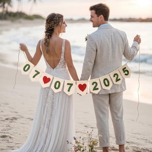 May include: A decorative banner featuring off-white fabric pennants strung together with twine. The pennants display the green felt numbers 07, 07, 2025 and two red crocheted heart shapes positioned between the date segments for a wedding display.