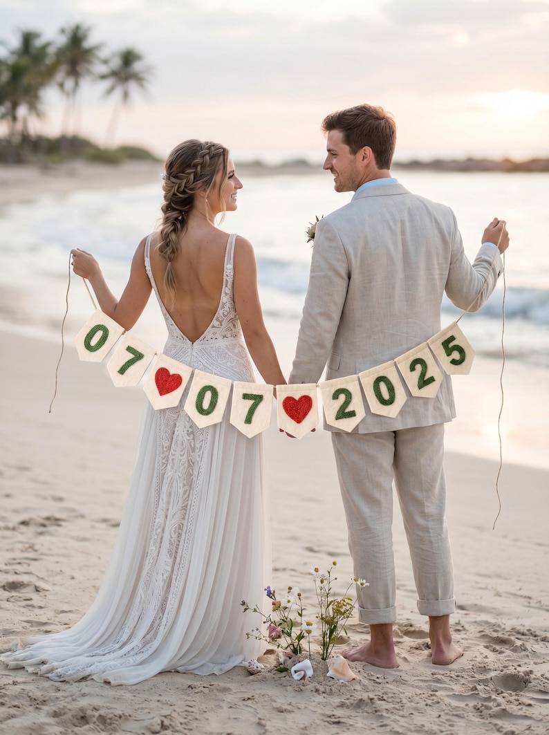May include: A decorative banner featuring off-white fabric pennants strung together with twine. The pennants display the green felt numbers 07, 07, 2025 and two red crocheted heart shapes positioned between the date segments for a wedding display.