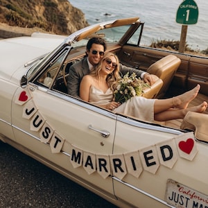 May include: A cream-coloured vintage convertible car decorated with a white pennant banner featuring the black text JUST MARRIED and red heart icons. The car has tan leather interior seating and a California 1 highway sign is visible nearby.