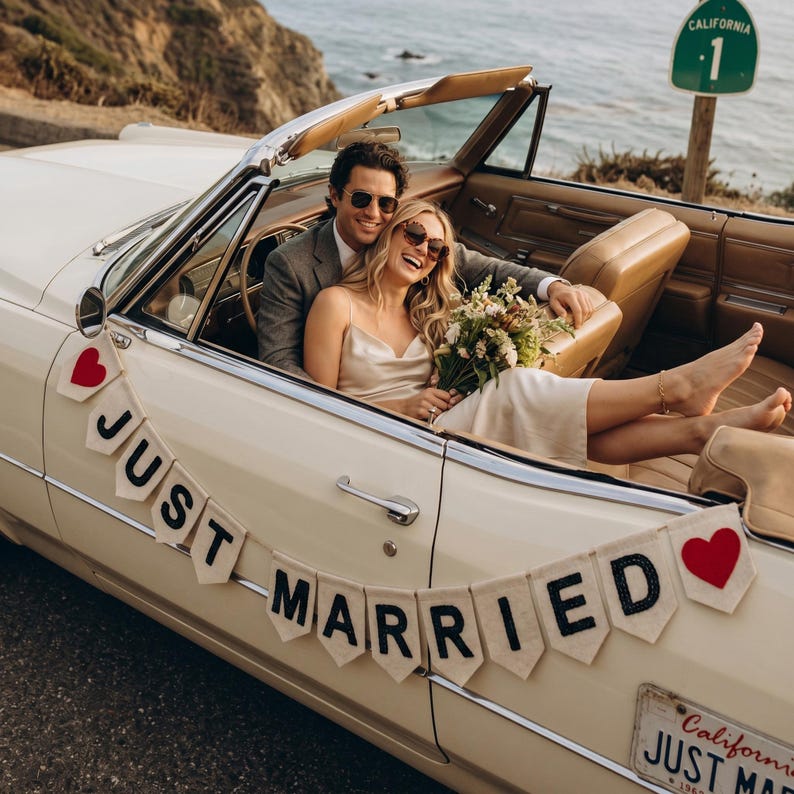 May include: A cream-coloured vintage convertible car decorated with a white pennant banner featuring the black text JUST MARRIED and red heart icons. The car has tan leather interior seating and a California 1 highway sign is visible nearby.