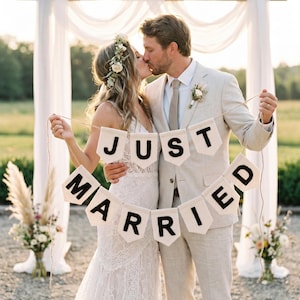 May include: A cream-coloured fabric banner featuring the black text "JUST MARRIED" printed on individual pennant-shaped flags strung together with twine. The banner consists of two rows of pennants, with "JUST" on the top and "MARRIED" on the bottom.