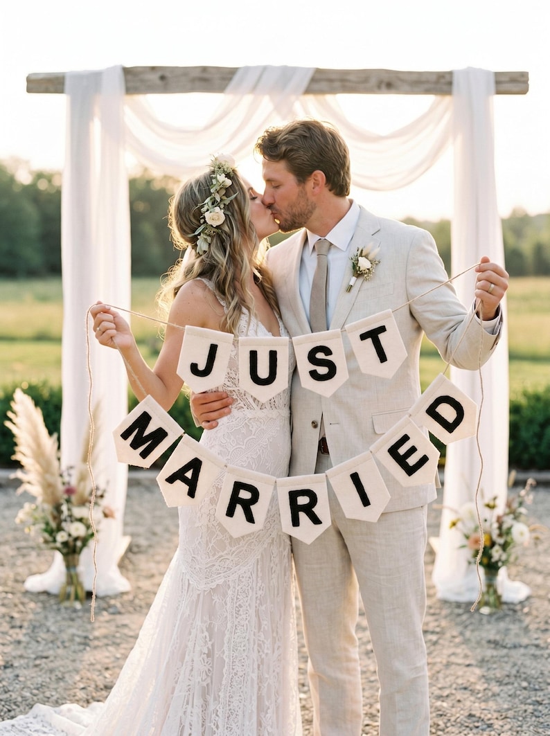 May include: A cream-coloured fabric banner featuring the black text "JUST MARRIED" printed on individual pennant-shaped flags strung together with twine. The banner consists of two rows of pennants, with "JUST" on the top and "MARRIED" on the bottom.