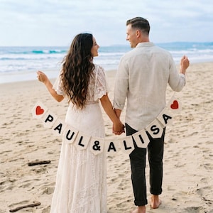 May include: A cream-coloured fabric banner featuring the text PAUL & ALISA in black capital letters, flanked by two red heart icons on each end. The banner is suspended by a thin string and held horizontally between two individuals on a sandy beach.